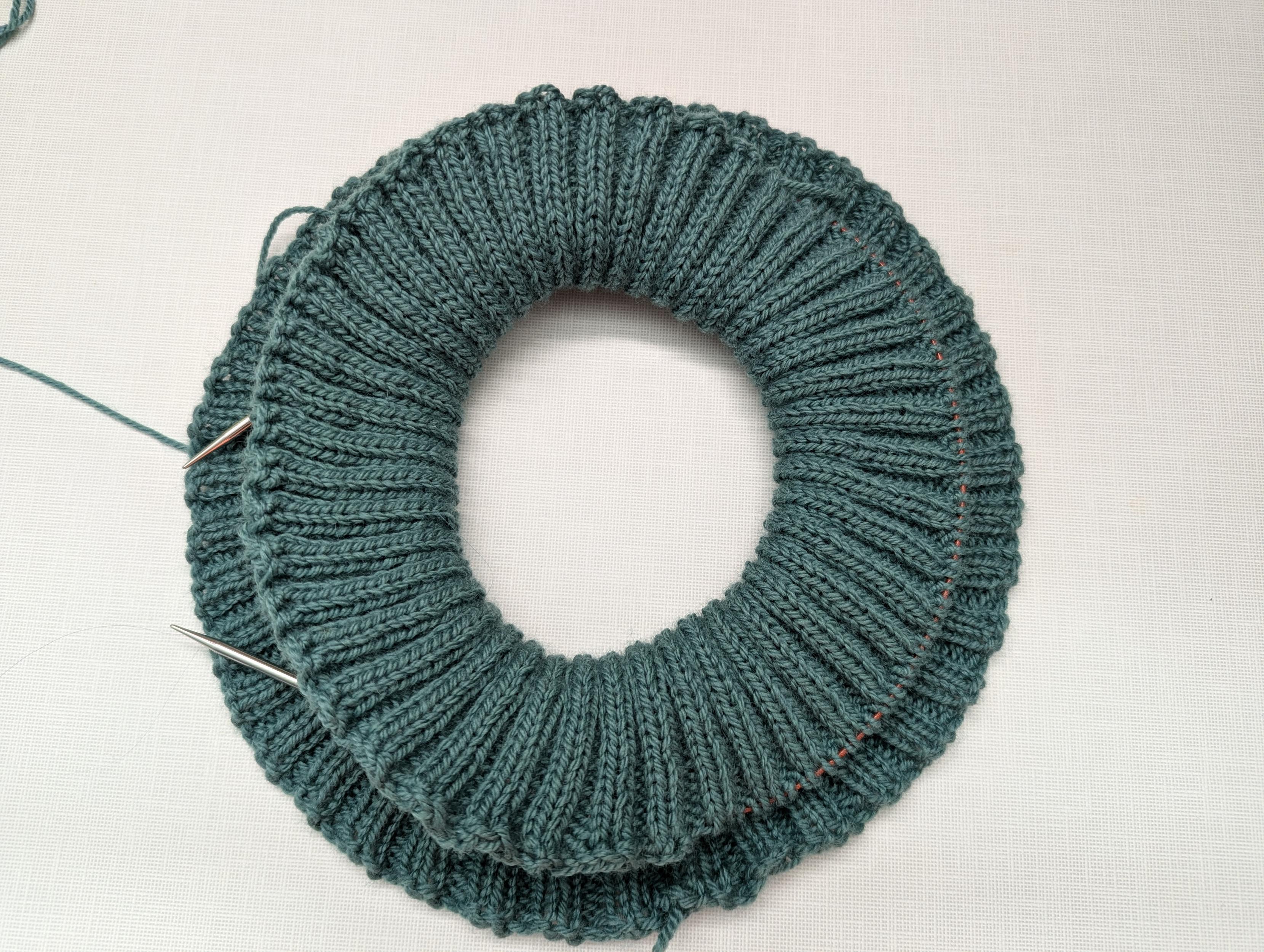 A ring of fabric resulting from a circular cuff knitted in a k2p2 pattern folding over itself in green yarn.  A circular needle runs through about 1/3rd of the top of the cuff and the rest of the cuff has been bound off