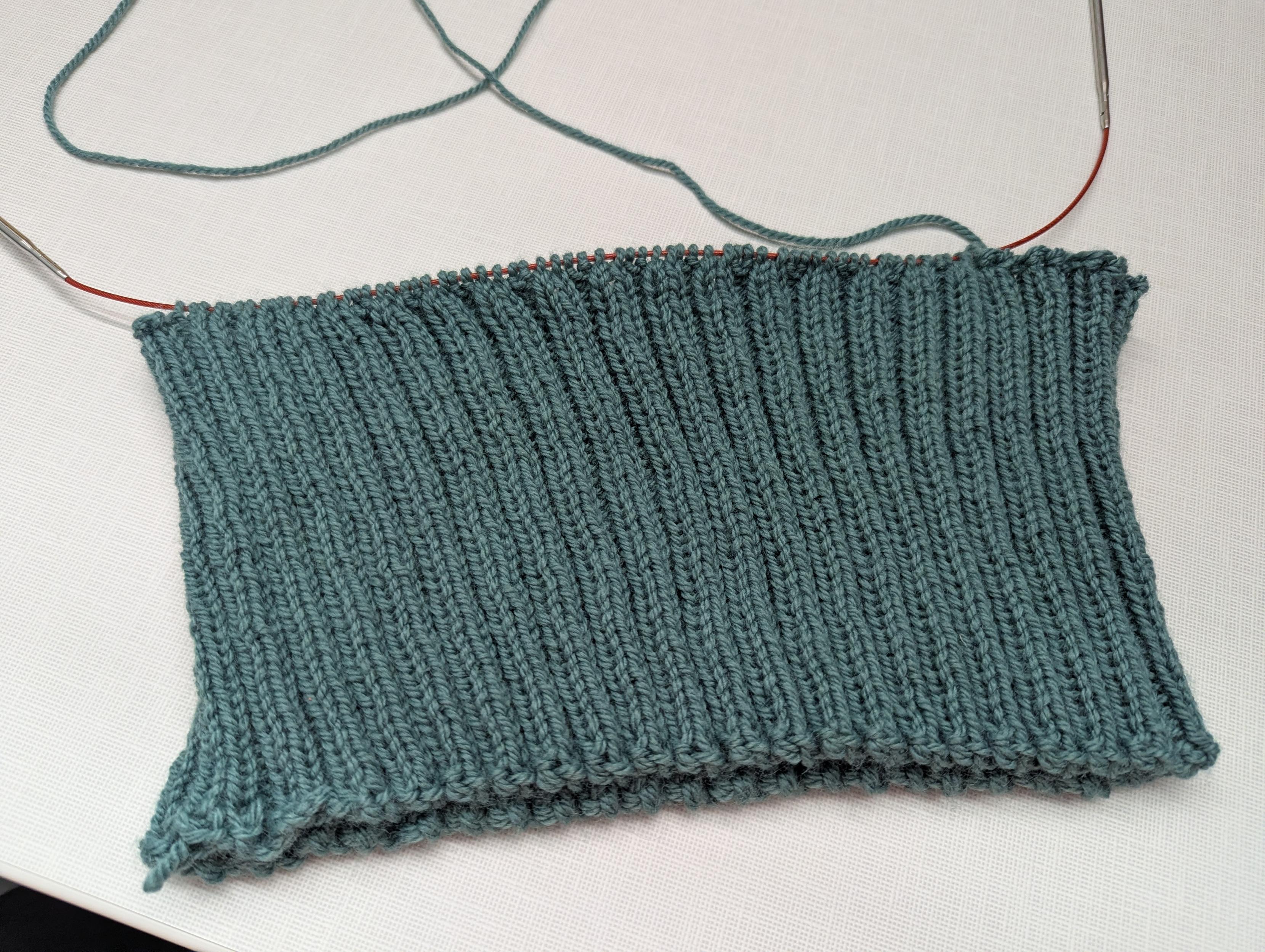 A circular cuff knitted in a k2p2 pattern made of green yarn that is a out 14 cm high.  A circular needle runs through about 1/3rd of the top of the cuff and the rest of the cuff has been bound off