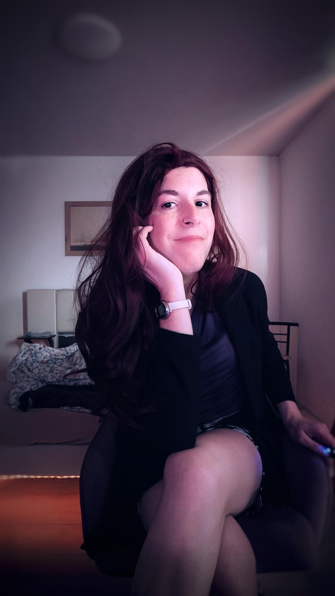 Miona sitting in a desk chair, looking at the camera. She wears a purple blouse, a black blazer, and a black midi skirt with pink and white flower print.